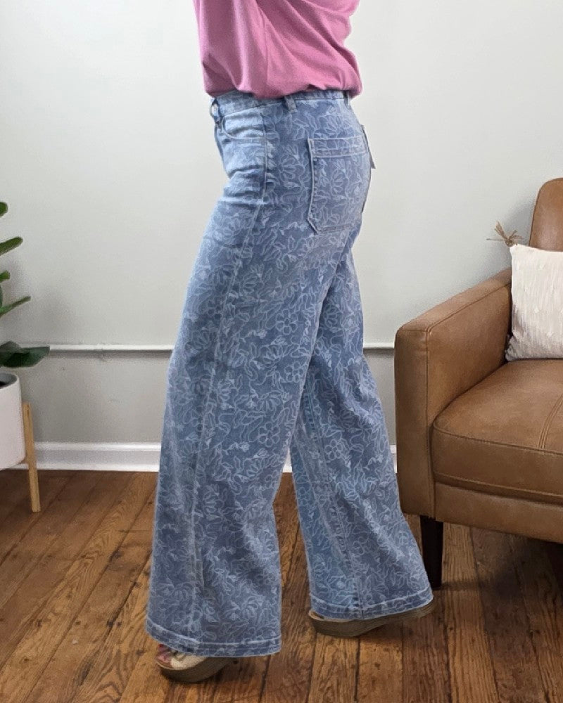A person wearing the Briella Floral Print, High Rise, Wide Leg Jeans from Vervet is standing on a wooden floor near a tan chair and plant against a white wall. Only the lower half of the person is visible.