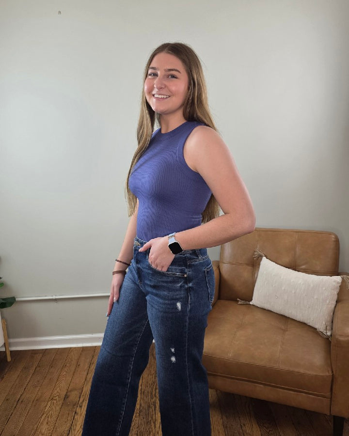 Woman standing in a living room wearing the Burke cropped tank top in the color Marlin and wide leg, dark wash jeans. 
