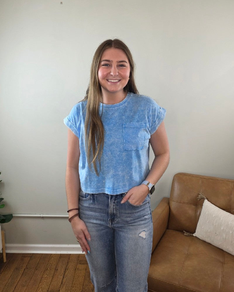 A young woman with long brown hair smiles at the camera indoors, wearing the ZENANA Candace Snow Washed Cropped Short Sleeve Top, ripped jeans, and a smartwatch. A brown couch and a plant can be seen in the background.