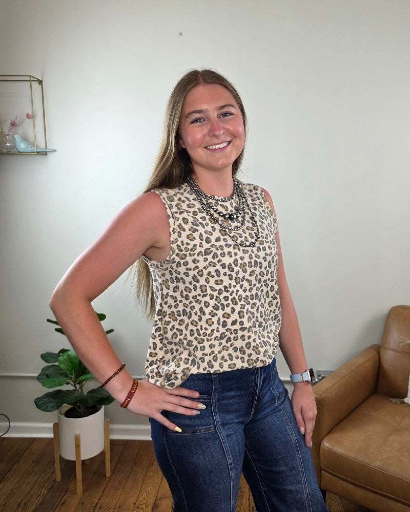 A woman with long brown hair, wearing the Haptics Cleo Leopard Print Tank Top and blue jeans, smiles in a living room featuring a tan chair, potted plant, and shelf in the background.