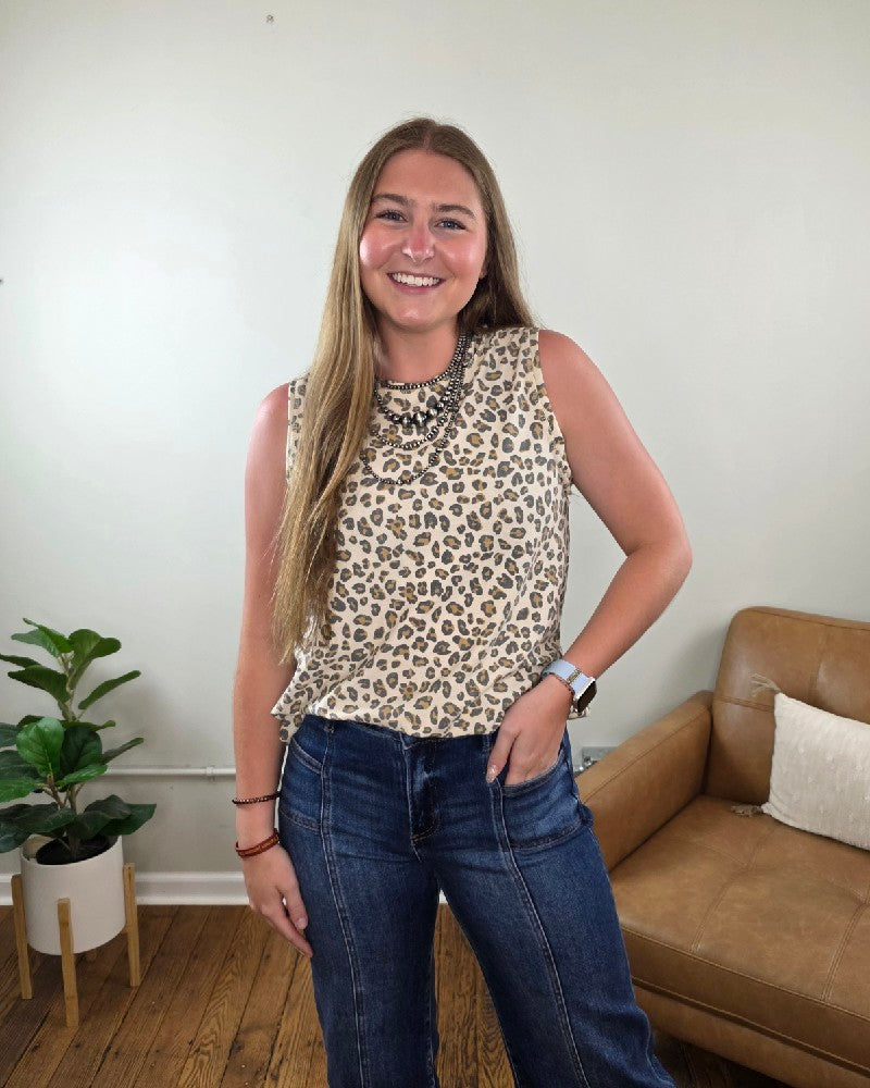 A woman with long hair smiles indoors, wearing the Haptics Cleo Leopard Print Tank Top and blue jeans, standing by a brown leather chair and a potted plant on a wooden floor.