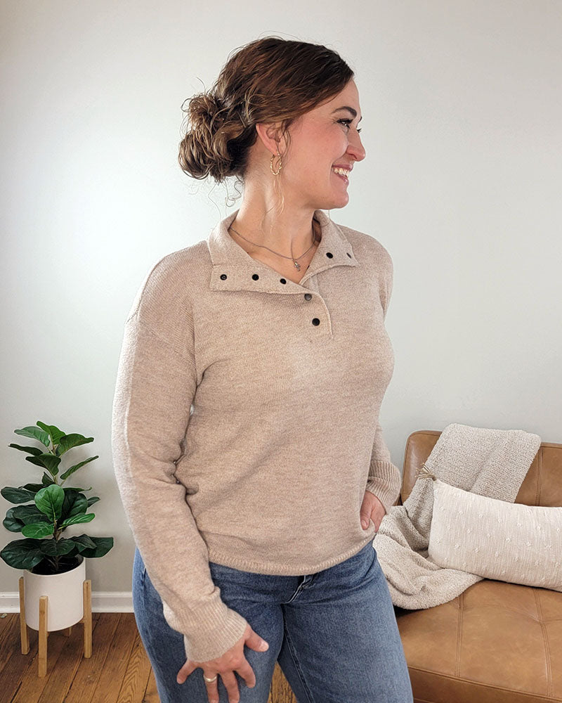A woman with brown hair in a bun smiles and looks left while wearing the Staccato Devon Half Snap Sweater and blue jeans, standing in a cozy living room with a tan couch, white blanket, and green potted plant.