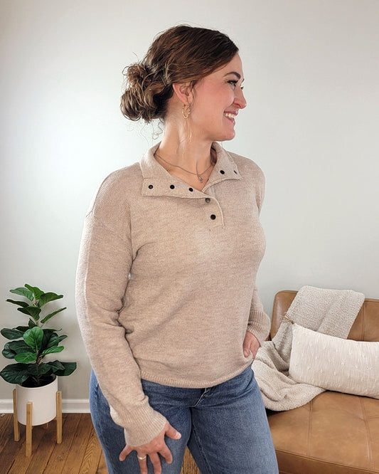 A woman with brown hair in a bun smiles and looks left while wearing the Staccato Devon Half Snap Sweater and blue jeans, standing in a cozy living room with a tan couch, white blanket, and green potted plant.