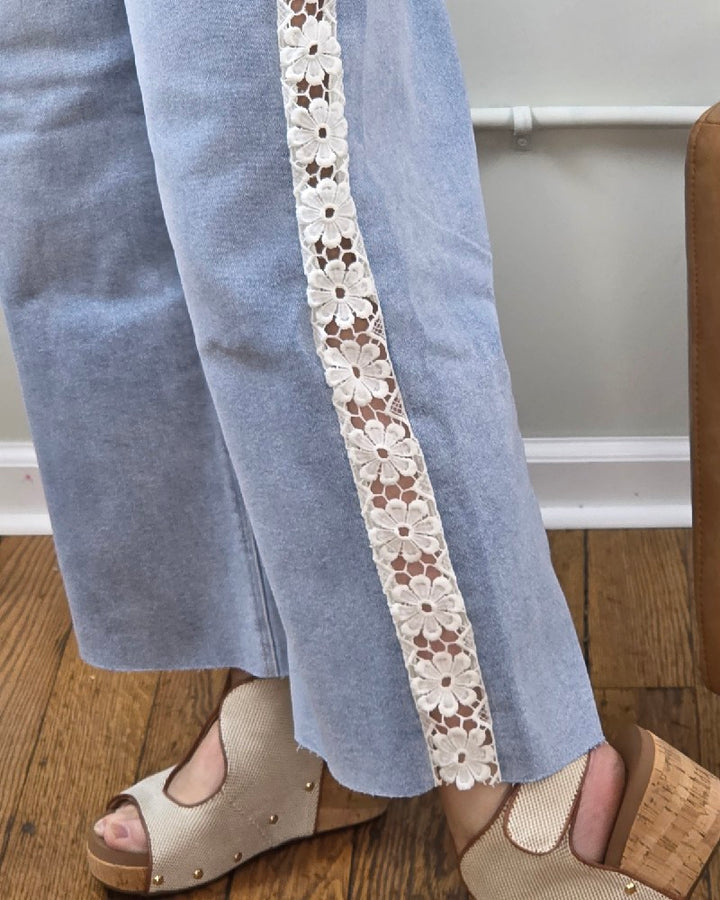 A person wears Risen Daisy Crop Wide Flower Lace Jeans—light-wash, high-rise, with a white floral lace inset—and beige open-toe sandals, standing on a wooden floor beside a tan bag.
