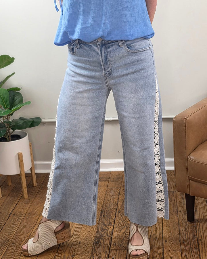 Someone stands on a wooden floor near a plant and brown chair, wearing Risen Daisy Crop Wide Flower Lace Jeans with white lace panels, a loose blue top, and beige wedge sandals.