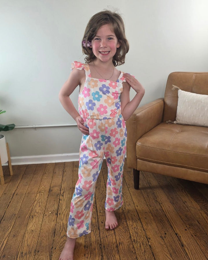 A young girl with shoulder-length brown hair smiles confidently, barefoot on a wooden floor in a bright room, wearing the Adorable Sweetness Kids Annabelle Floral Ruffle Jumpsuit with adjustable straps.