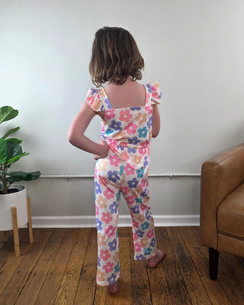 A young child with short brown hair stands indoors facing away, wearing the Adorable Sweetness Kids Annabelle Floral Ruffle Jumpsuit. Nearby are a potted plant and a brown armchair on the wooden floor.