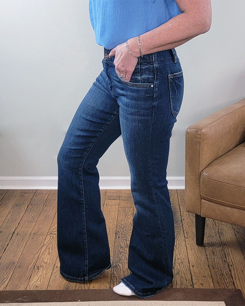 A person, shown from the waist down, stands barefoot except for a white sock on one foot while wearing Risen's Eleanor Flare Jeans. They are on wooden flooring next to a tan armchair.