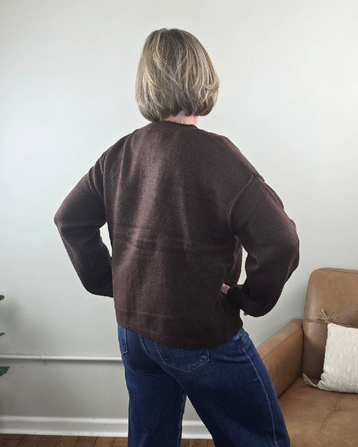 A person with short hair, wearing the Elma Button Down Loose Fit Sweater by HYFVE, Inc and blue jeans, stands with hands on hips facing away from the camera in a living room with a tan chair.