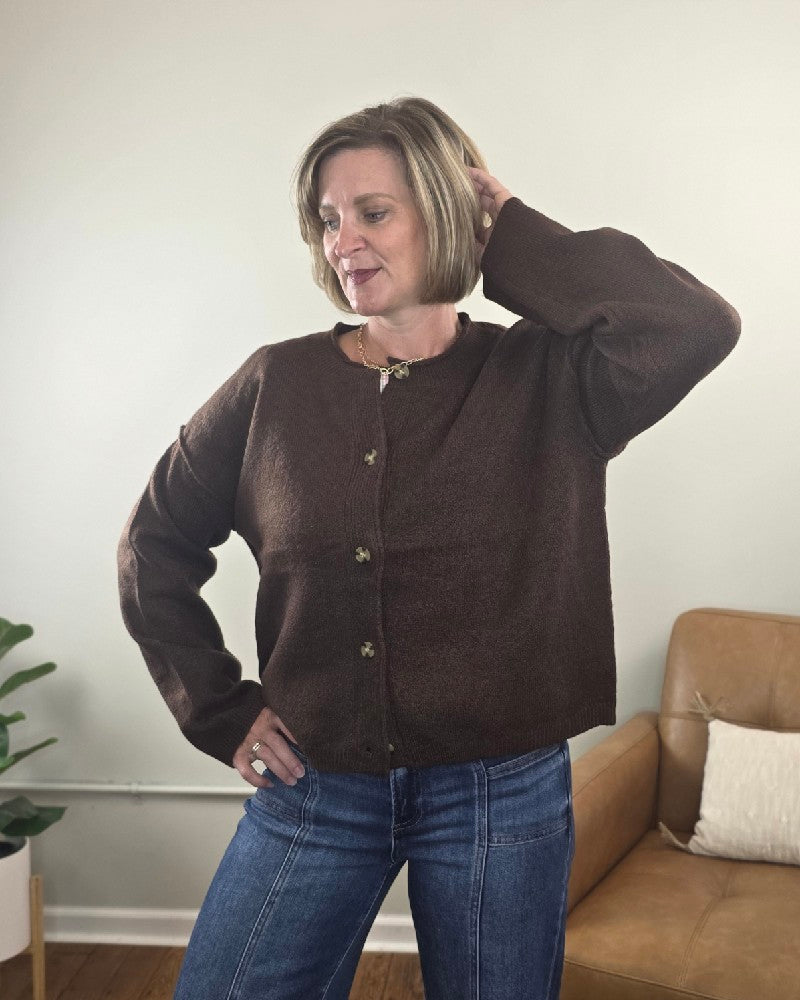 A woman with short blonde hair models the Elma Button Down Loose Fit Sweater by HYFVE, Inc, styled in brown with blue jeans. She stands in a cozy, modern living room, one hand on her hip and the other behind her head.
