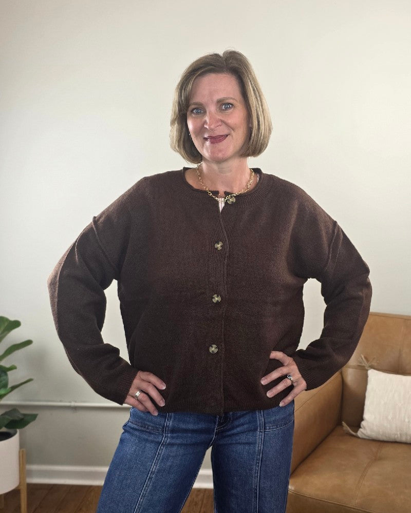 A woman with short blonde hair is standing indoors with her hands on her hips and smiling, wearing the HYFVE, Inc Elma Button Down Loose Fit Sweater in brown paired with blue jeans. A plant and a tan sofa are visible in the background.