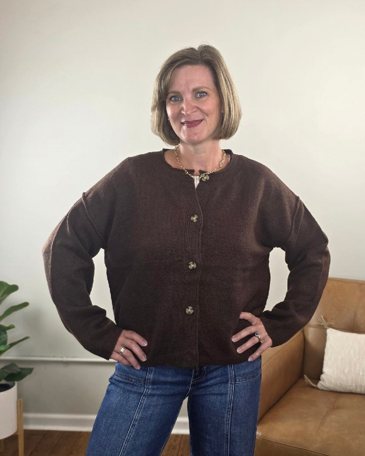 A woman with short blonde hair is standing indoors with her hands on her hips and smiling, wearing the HYFVE, Inc Elma Button Down Loose Fit Sweater in brown paired with blue jeans. A plant and a tan sofa are visible in the background.