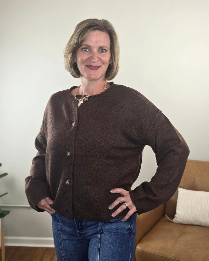 A woman with short hair stands indoors, smiling with one hand on her hip, wearing the Elma Button Down Loose Fit Sweater by HYFVE, Inc and blue jeans. In the background, there’s a couch and a plant.