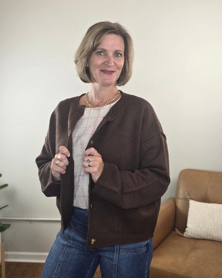 A woman with short hair, wearing the HYFVE, Inc Elma Button Down Loose Fit Sweater and blue jeans, stands indoors near a tan couch and a plant, smiling at the camera.