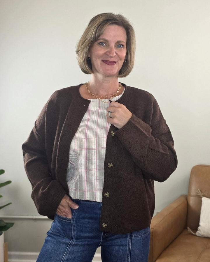 A woman with short light brown hair smiles at the camera, wearing the HYFVE, Inc Elma Button Down Loose Fit Sweater over a patterned blouse and blue jeans. She stands indoors near a tan couch and a green plant.