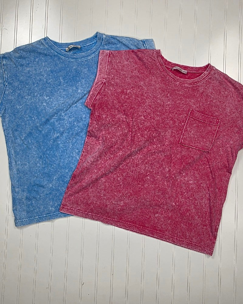 Two ZENANA Emma Snow Washed Short Sleeve Tops in 100% cotton are laid flat on a white wood surface: one blue without a pocket, the other red with a front chest pocket, both featuring a relaxed fit.