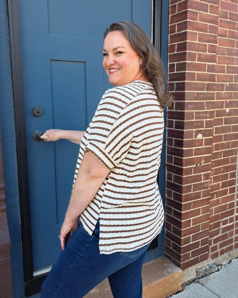 A woman with light brown hair, smiling by a blue door and red brick wall, wears the ZENANA Esther Striped Short Sleeve Hi-low Tee as a casual dress over blue jeans.