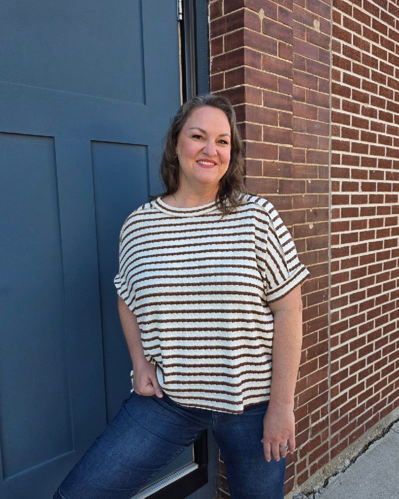 A woman with wavy brown hair smiles beside a blue door and red brick wall, wearing the ZENANA Esther Striped Short Sleeve Hi-low Tee with jeans and one hand in her pocket.