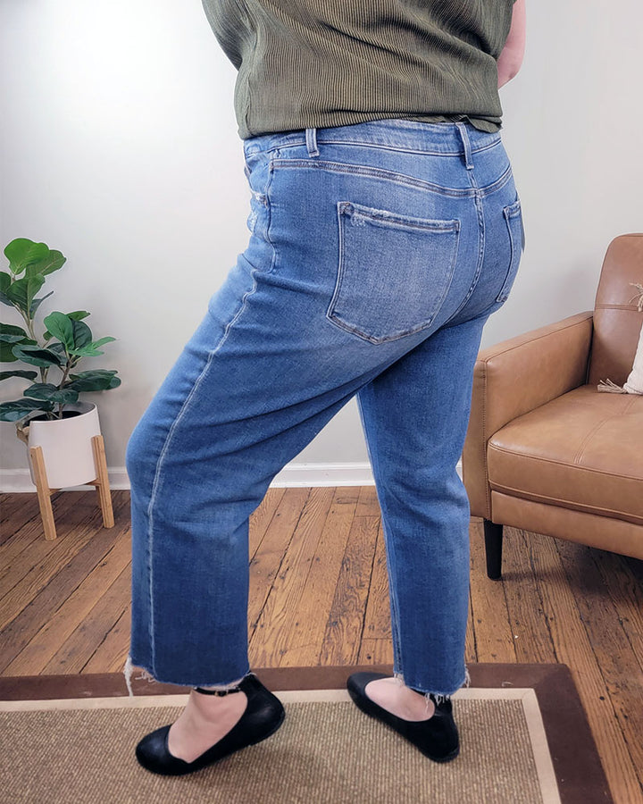 A person wearing Lovervet’s Kendall Straight Crop Jeans stands on a rug in a room with wooden floors, a potted plant, and a tan armchair. The back view highlights the jeans.