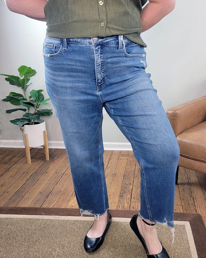 Wearing Lovervet’s Kendall Straight Crop Jeans, a person stands indoors on hardwood floors beside a potted plant and brown armchair, paired with black flats and a green striped shirt.