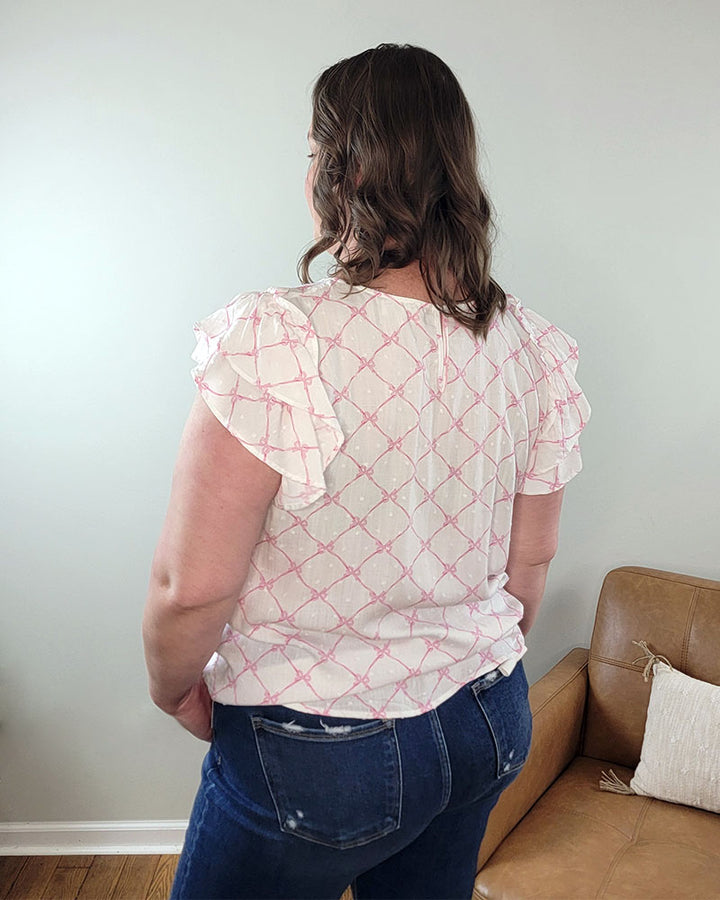 A woman with wavy brown hair stands with her back to the camera, wearing the Maddy Bow Top by Mine and E&M—a spring-summer blouse with a pink geometric pattern and ruffle sleeves—paired with blue jeans in a room featuring a tan couch.