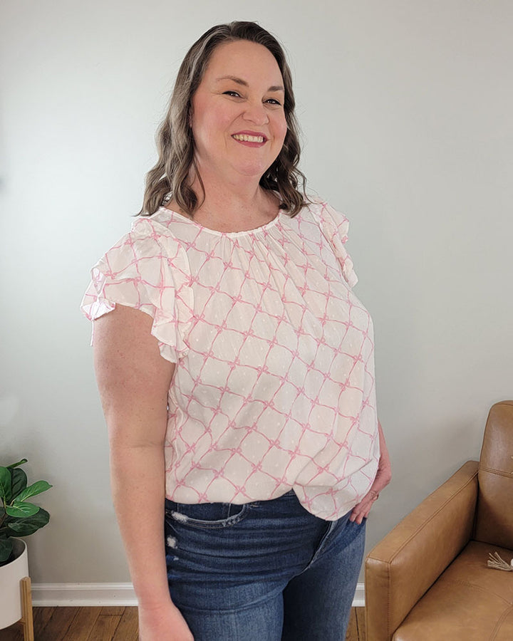 A woman with wavy brown hair smiles indoors, standing by a tan chair and green potted plant. She wears the Mine and E&M Maddy Bow Top, a spring-summer blouse with pink patterns, paired with blue jeans against a light wall.