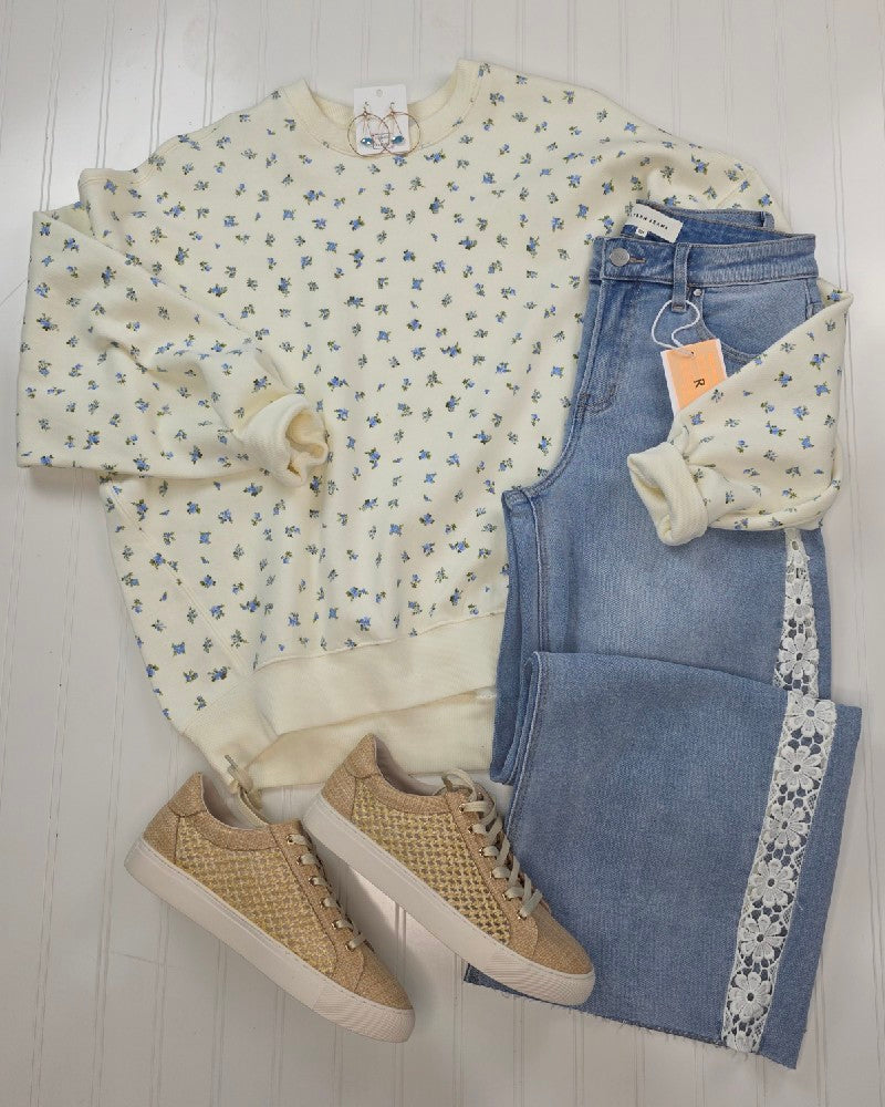 The HYFVE, Inc Fiona Floral Print Crewneck, with delicate blue flowers, is styled with light-wash jeans featuring white lace trim and tan woven sneakers, all arranged on a white wooden floor for a casual look.