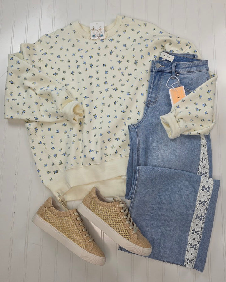The HYFVE, Inc Fiona Floral Print Crewneck, with delicate blue flowers, is styled with light-wash jeans featuring white lace trim and tan woven sneakers, all arranged on a white wooden floor for a casual look.