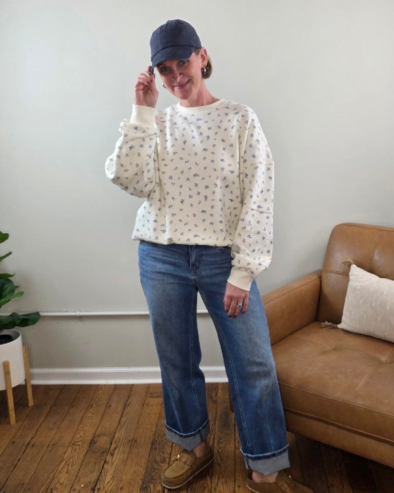 A person stands indoors by a tan couch and potted plant, smiling in the HYFVE, Inc Fiona Floral Print Crewneck, loose blue cuffed jeans, tan loafers, and a navy cap for a relaxed casual look.