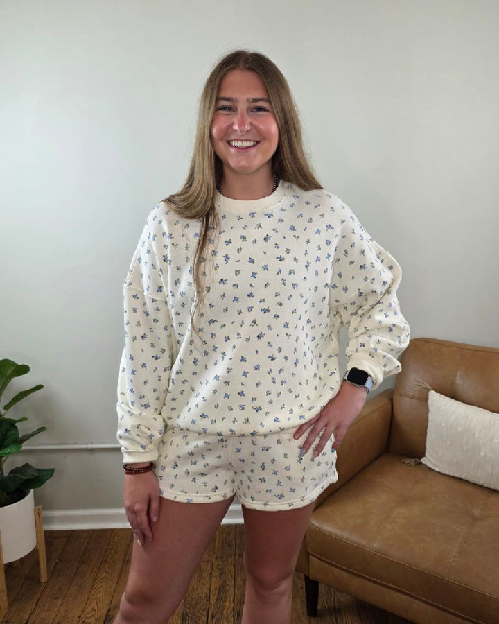 A woman with long light brown hair smiles indoors next to a tan couch and plant, wearing the Fiona Floral Print Crewneck by HYFVE, Inc and matching shorts, both in cream with a small blue pattern.