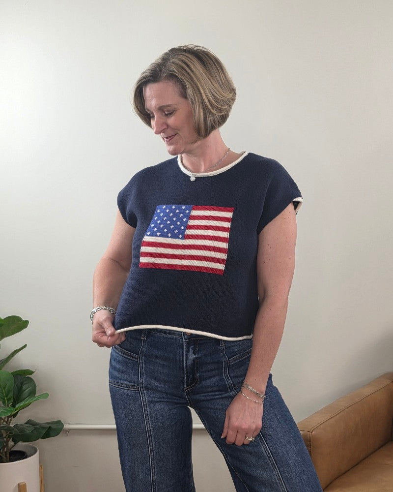 A woman with short blonde hair stands indoors by a tan couch and green plant, looking down and holding the hem of her HYFVE, Inc Flag Knit Short Sleeve Top, paired with blue jeans.