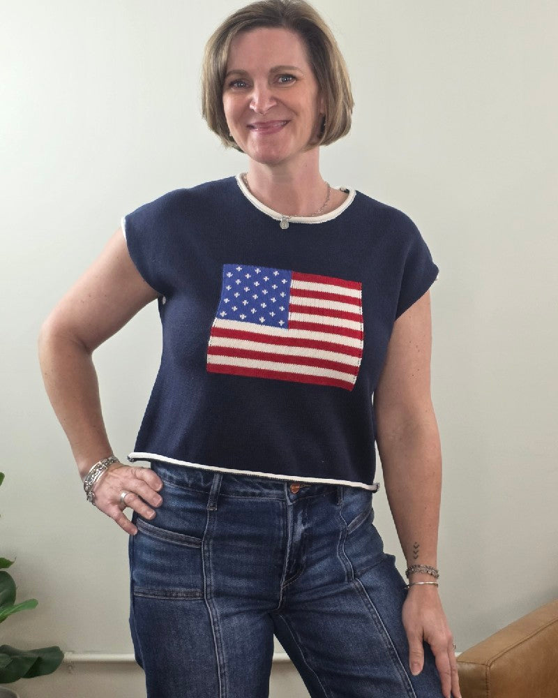 A woman with short hair wears the HYFVE, Inc Flag Knit Short Sleeve Top, featuring an American flag, paired with blue jeans. She stands indoors, smiling confidently with one hand on her hip.
