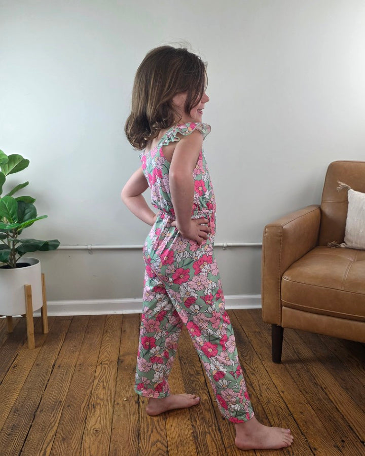 A young girl stands in profile on a wooden floor, smiling with her hands on her hips. She wears the Kids Kassidy Floral Ruffle Jumpsuit by Adorable Sweetness. A tan armchair and a potted plant can be seen in the background.