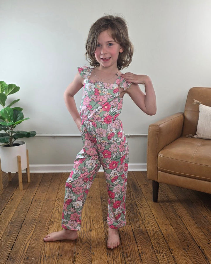 A young child stands barefoot on a wooden floor in the Adorable Sweetness Kids Kassidy Floral Ruffle Jumpsuit with adjustable straps, near a light brown armchair and green potted plant against a plain, light-colored wall.