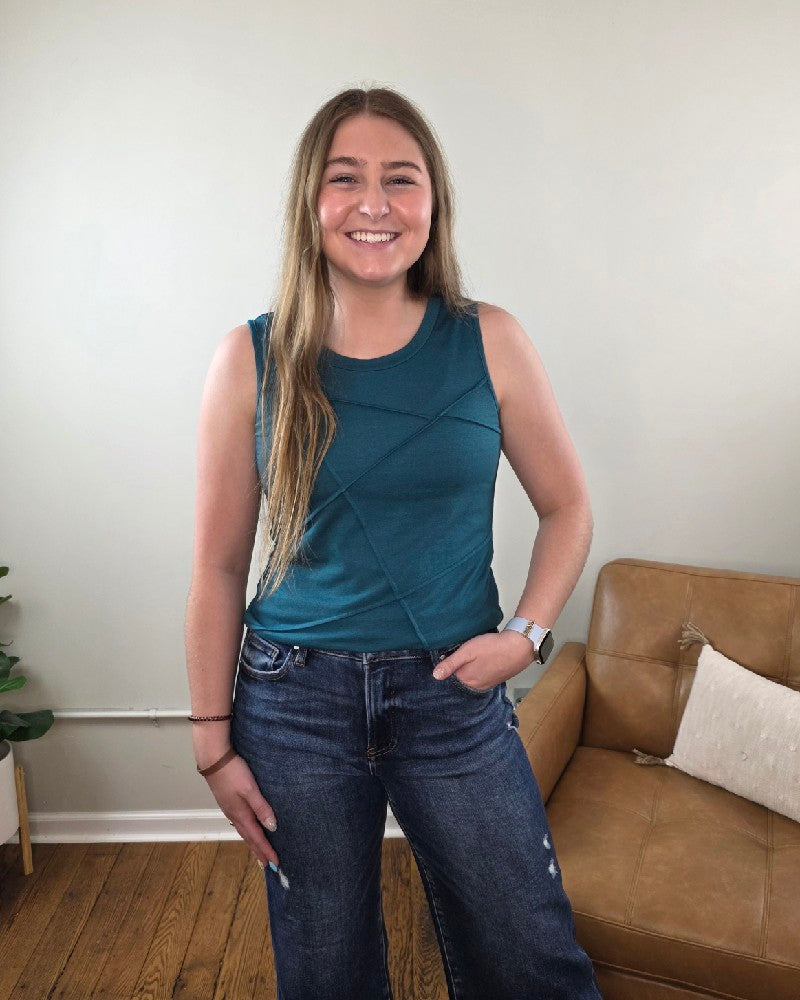 A smiling woman with long hair stands indoors, wearing the Lovesoft Freya Tank Top With Stitching Details and jeans. She has one hand in her pocket and stands by a tan leather chair against a light-colored wall.