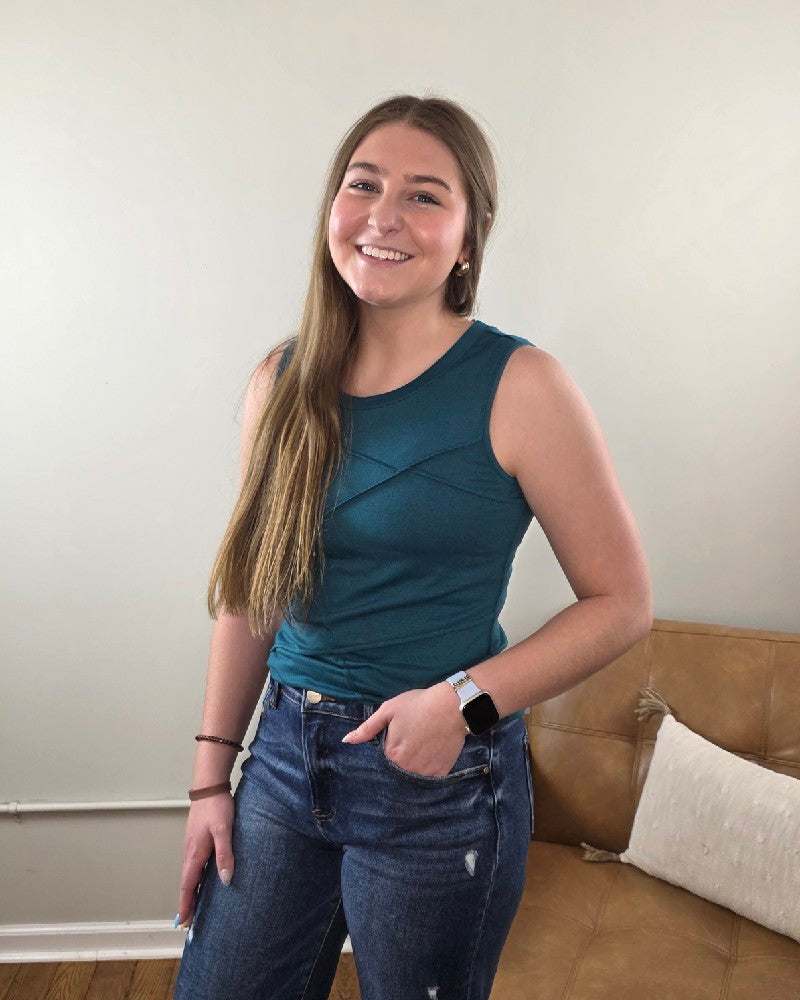 A young woman with long brown hair, wearing a classic Lovesoft Freya Tank Top With Stitching Details and blue jeans, smiles in a room with a tan couch and white pillow in the background.