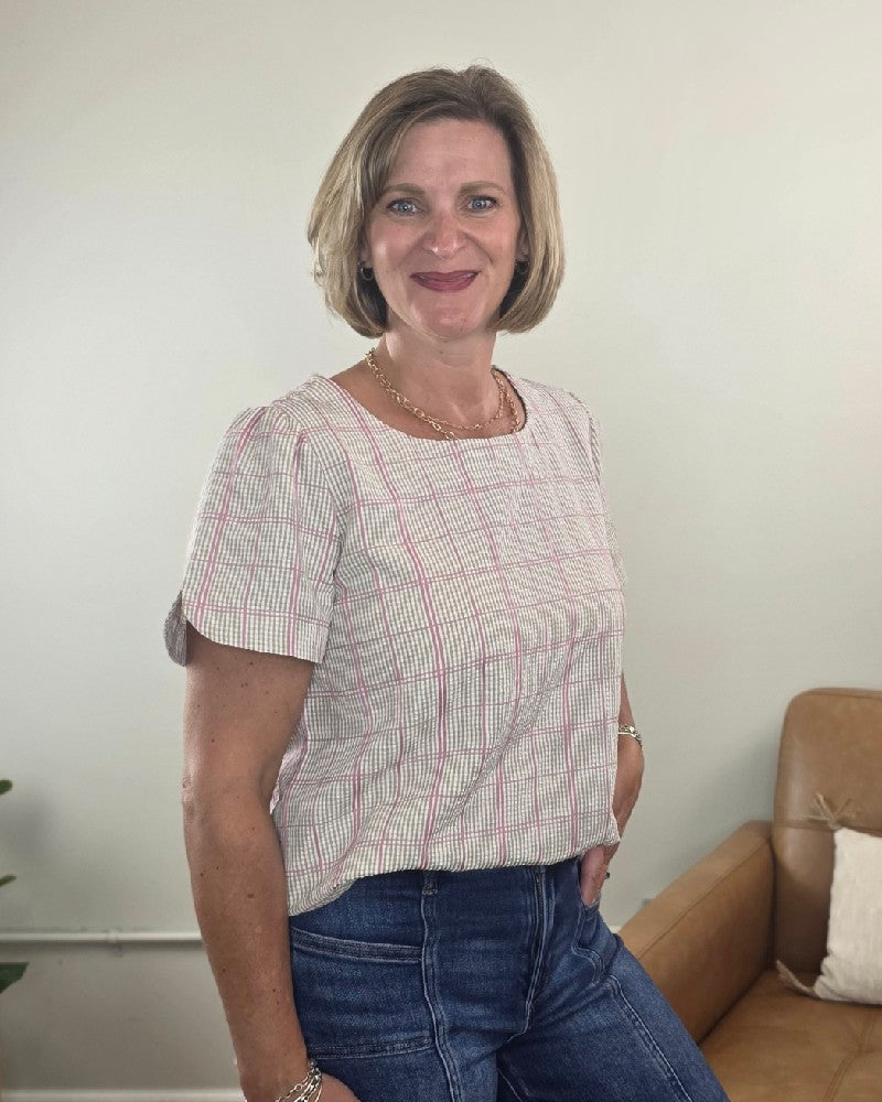 A woman with short blond hair, wearing the Les Amis Georgina Khaki Plaid Blouse—a lightweight, plaid transitional blouse—smiles beside a tan couch in a well-lit room.