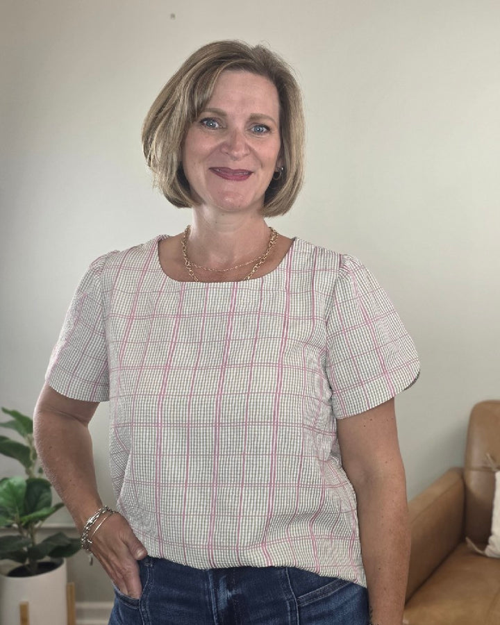 A woman with short blonde hair, wearing the Les Amis Georgina Khaki Plaid Blouse and jeans, stands indoors smiling. In the background, there is a plant and a tan chair set against a plain wall.