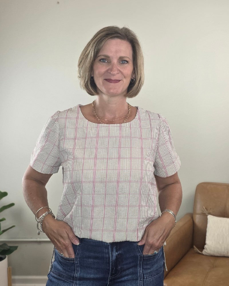 A woman with short blonde hair stands indoors smiling with her hands in her pockets, wearing the Les Amis Georgina Khaki Plaid Blouse and jeans. A green plant and a tan couch are visible in the background.