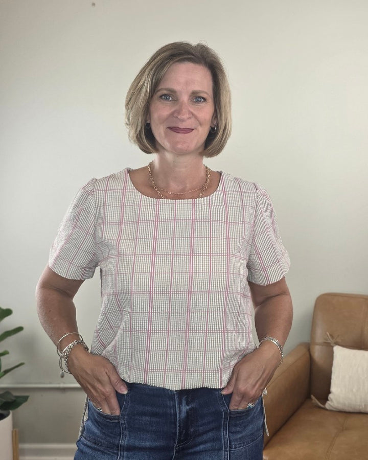 A woman with short blonde hair stands indoors smiling with her hands in her pockets, wearing the Les Amis Georgina Khaki Plaid Blouse and jeans. A green plant and a tan couch are visible in the background.