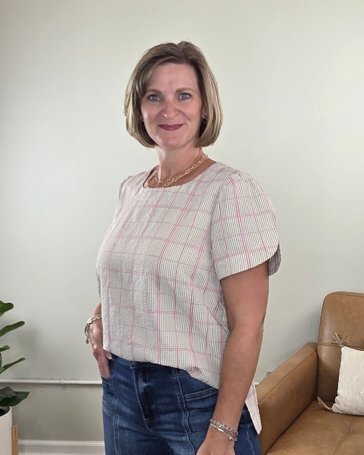 Wearing the Les Amis Georgina Khaki Plaid Blouse, a woman with short hair stands indoors by a tan couch and a green plant, smiling at the camera.