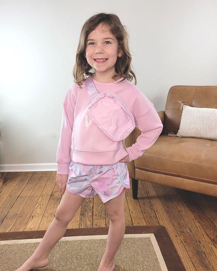 A smiling young girl stands indoors on a rug, wearing the Reflex Little Girls Shay Crewneck Shorts and Bag Set in pink. A tan couch with a white pillow sits behind her on wooden flooring.