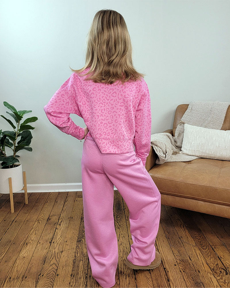 A person with shoulder-length blonde hair faces away, wearing the Reflex Girls Jessi Leopard Set in pink, made from lightweight scuba fabric. They stand in a room with wooden floors, a tan couch, and a potted plant.