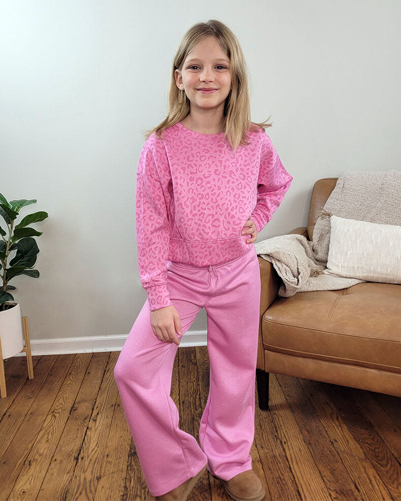 A young girl stands indoors on a wooden floor, smiling with one hand on her hip. She wears the Reflex Girls Jessi Leopard Set, featuring a leopard-print pullover and wide-leg pants. Behind her is a tan couch with cream blankets and a green potted plant.