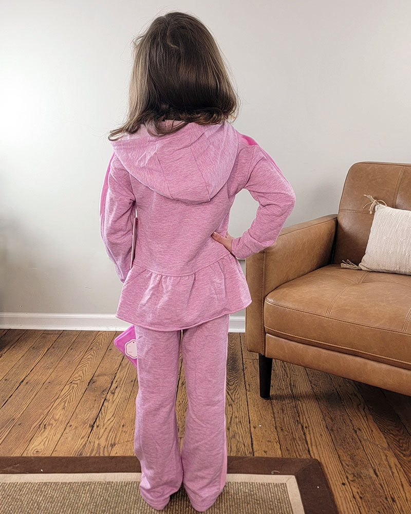 A young girl with brown hair faces away from the camera, wearing the Reflex Little Girls Lena Hoodie + Pant Set. She stands on a wooden floor beside a tan armchair with a white pillow.