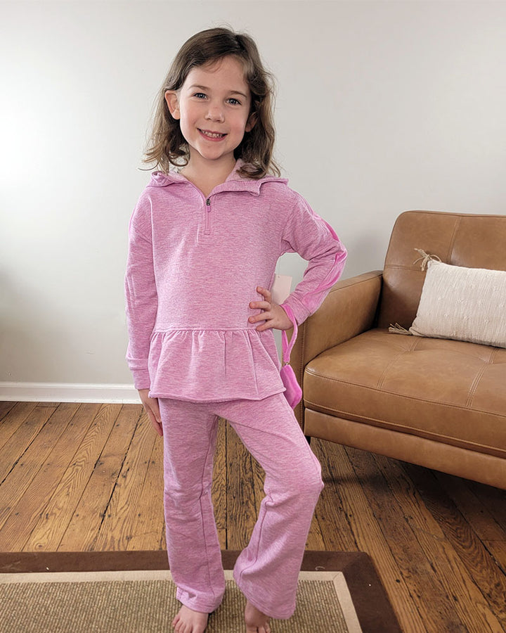 A young girl smiles indoors, wearing the Reflex Little Girls Lena Hoodie + Pant Set. She poses with one hand on her hip in front of a brown couch and white wall, standing on a wooden floor.