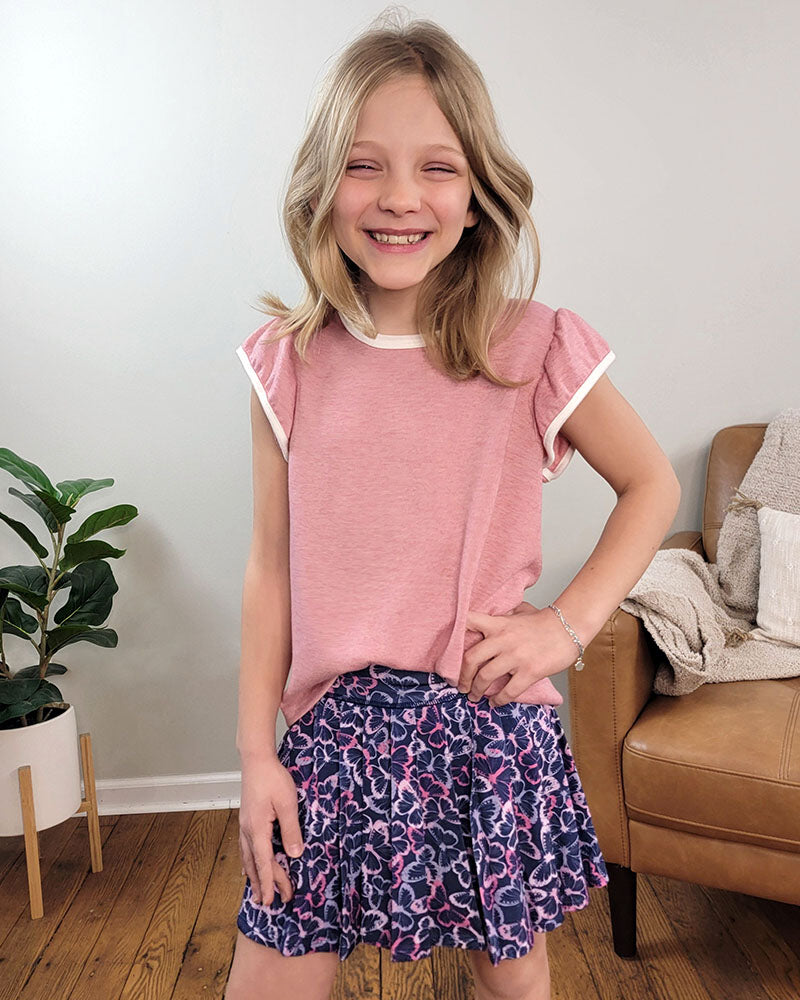 A smiling young blonde girl wears the Reflex Girls Lydia Butterfly Skort with a pink shirt, standing indoors by a brown chair and green plant on a wooden floor.