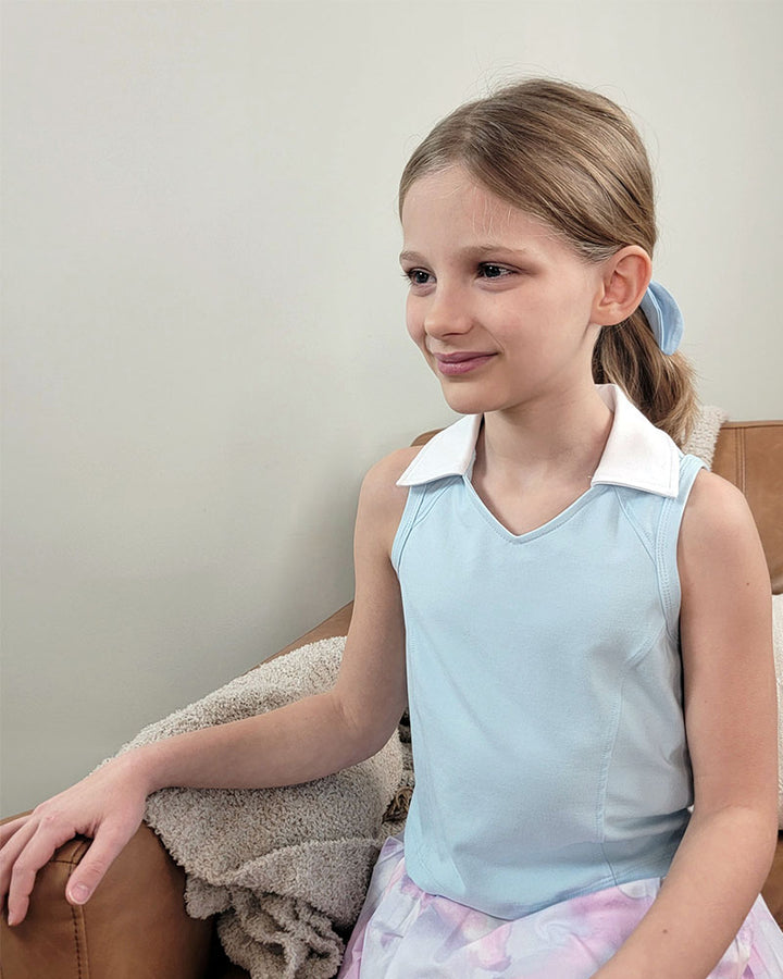 A young girl with light brown hair tied back in a blue ribbon smiles on a tan couch, wearing the Reflex Girls Nyla Tank and Skort Set. A beige blanket beside her completes her charming and comfortable look.