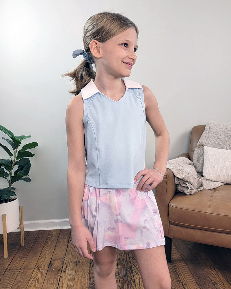 A young girl smiles indoors wearing the Reflex Girls Nyla Tank and Skort Set in light blue, pastel pink, and purple. Behind her are a tan couch with pillows and a white potted plant.