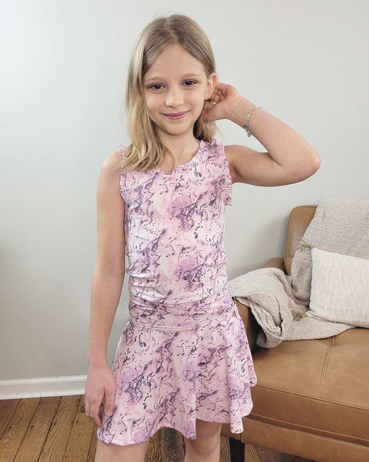 A young blonde girl smiles indoors, touching her hair while wearing the Reflex Girls Tate Tank Top with Ruffle Details and Matching Skort Set in a pink and purple marble splatter design. She stands by a tan couch decorated with a blanket and pillows.